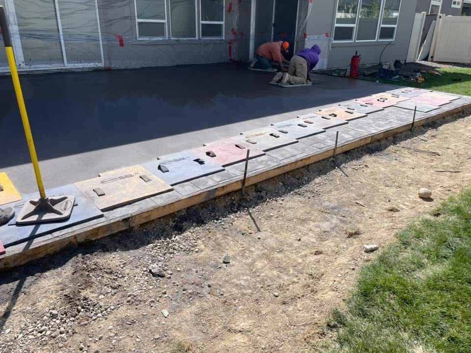 Workers installing paver stones along a building wall, with a gravel strip and grassy edge nearby