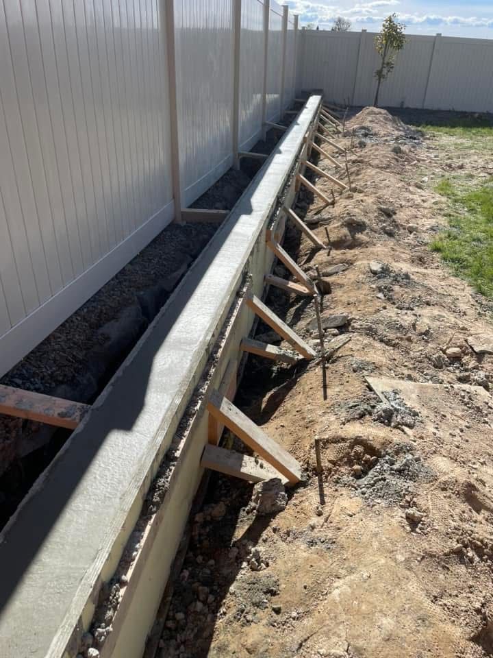 Concrete footing beside a vinyl fence, with wooden braces supporting fresh cement along a narrow trench.
