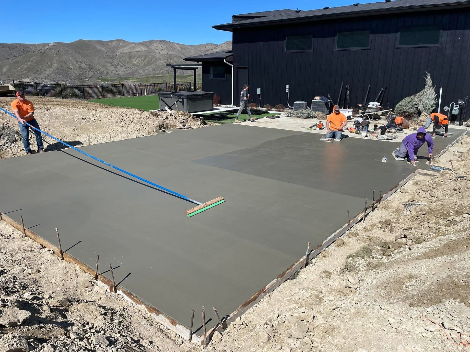 Workers smoothing fresh concrete on a large patio slab beside a dark building in a desert setting