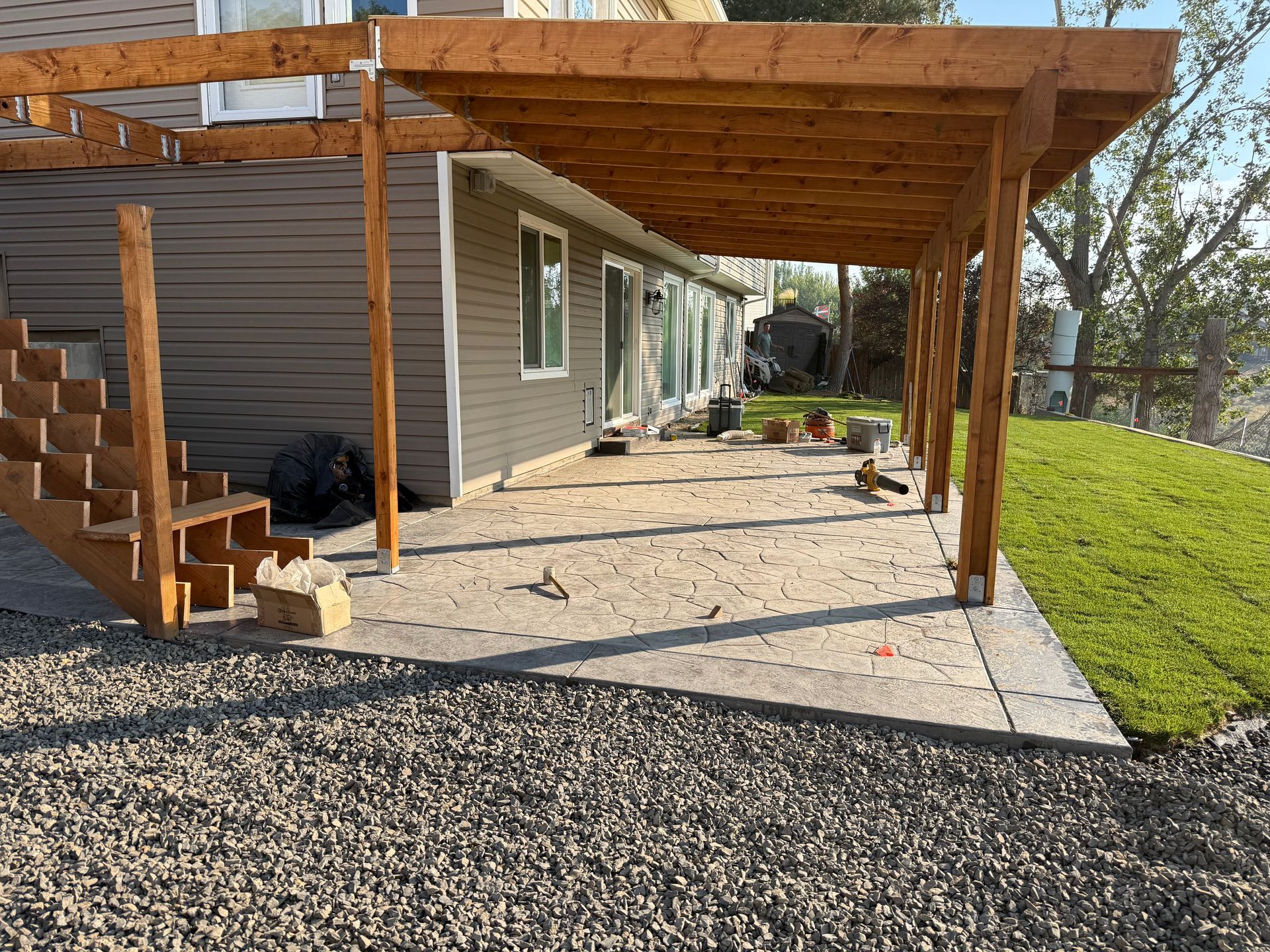 Covered patio with wooden pergola beside a house, concrete pad, stairs, and gravel yard
