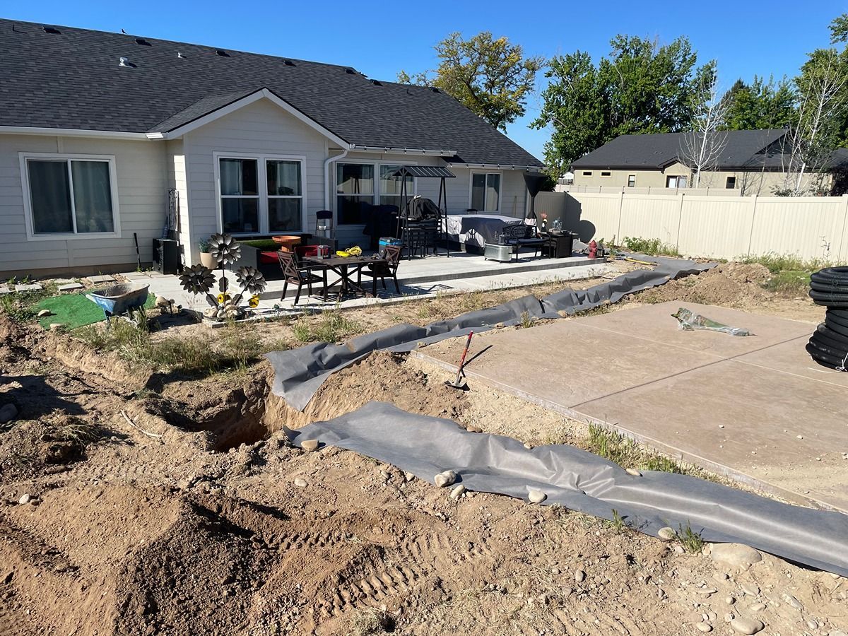 Backyard under construction beside a house, with dug-up soil, stone borders, and covered patio area