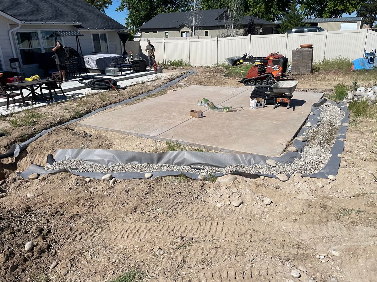 Backyard construction site with a leveled dirt pad, tarp edging, and a wheelbarrow near a fenced yard.