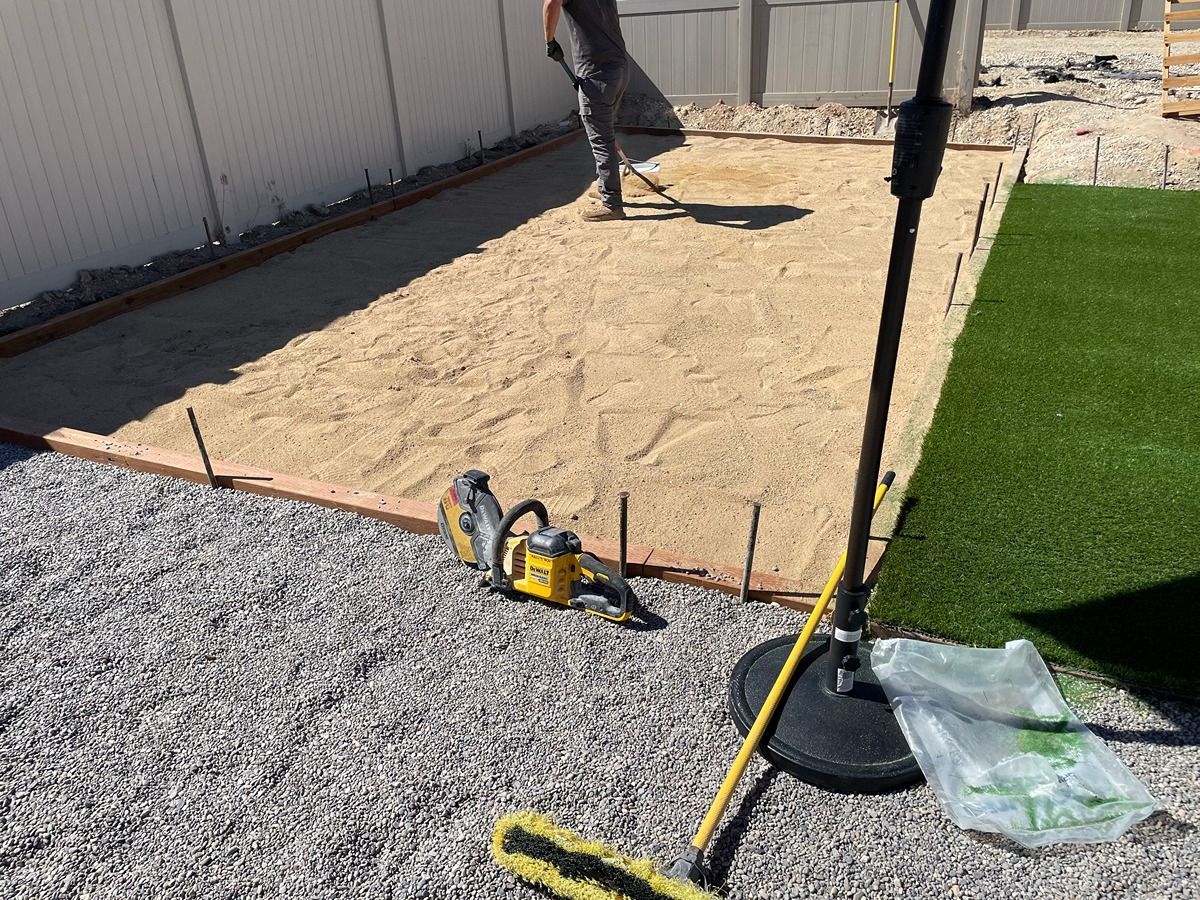 Backyard patio under construction, with gravel, sand, and a worker using a shovel