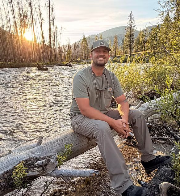 Man sitting on a log beside a river at sunset, with mountains and autumn trees in the background