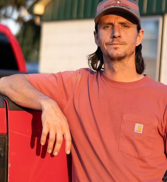 Man leaning on a red truck, wearing a salmon shirt and cap, outdoors in warm sunlight