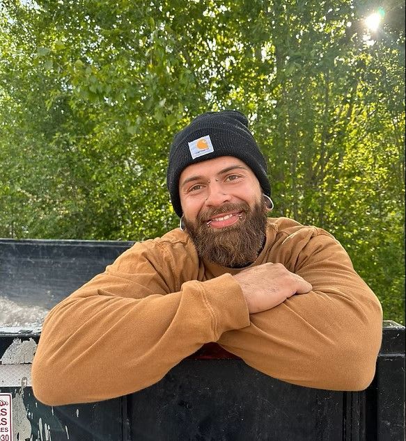 Smiling man in a black beanie and tan hoodie with folded arms outdoors in front of green trees