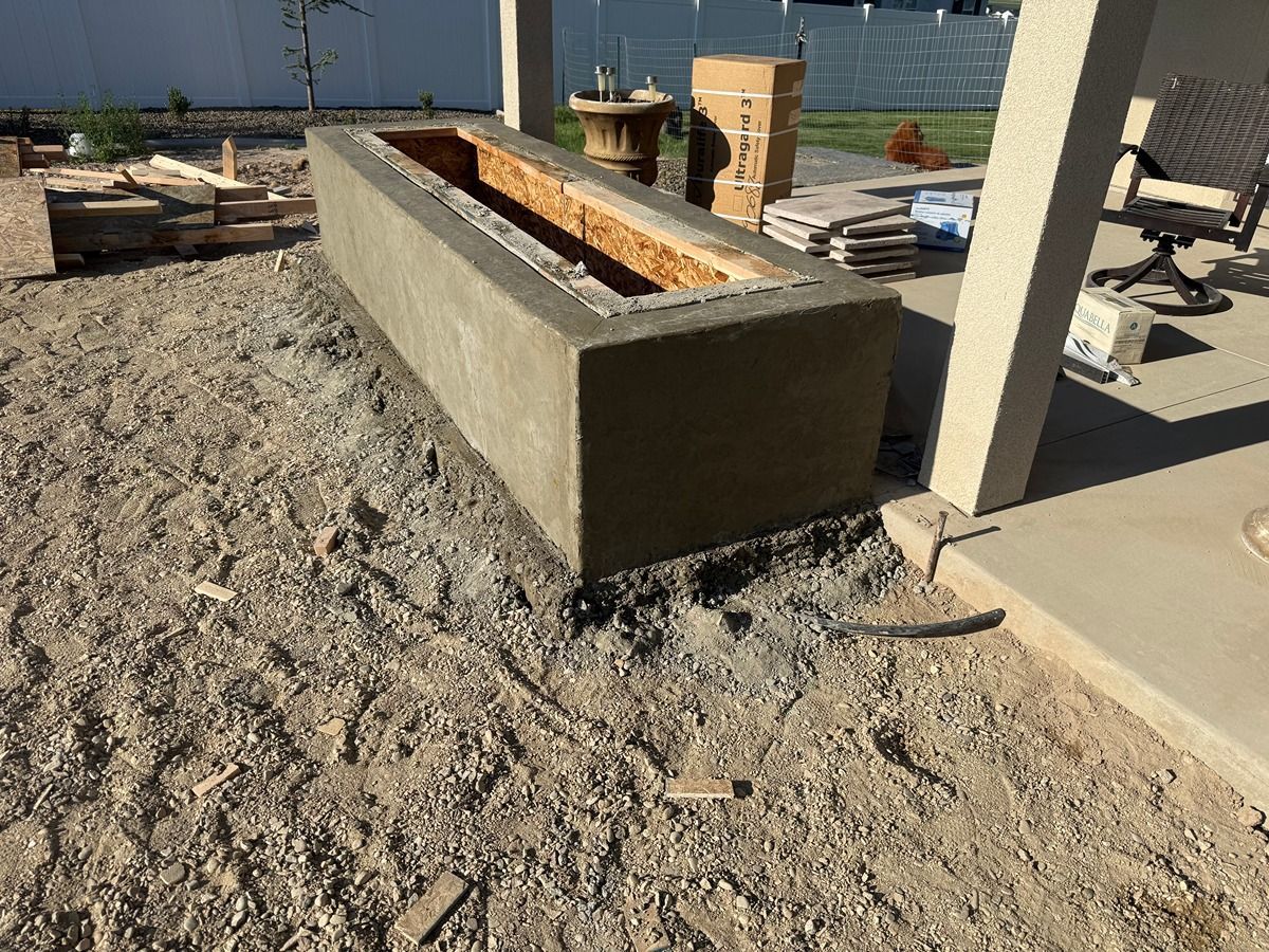Outdoor concrete fire pit beside a patio in a gravel yard, with stacked bricks and a fence in the background