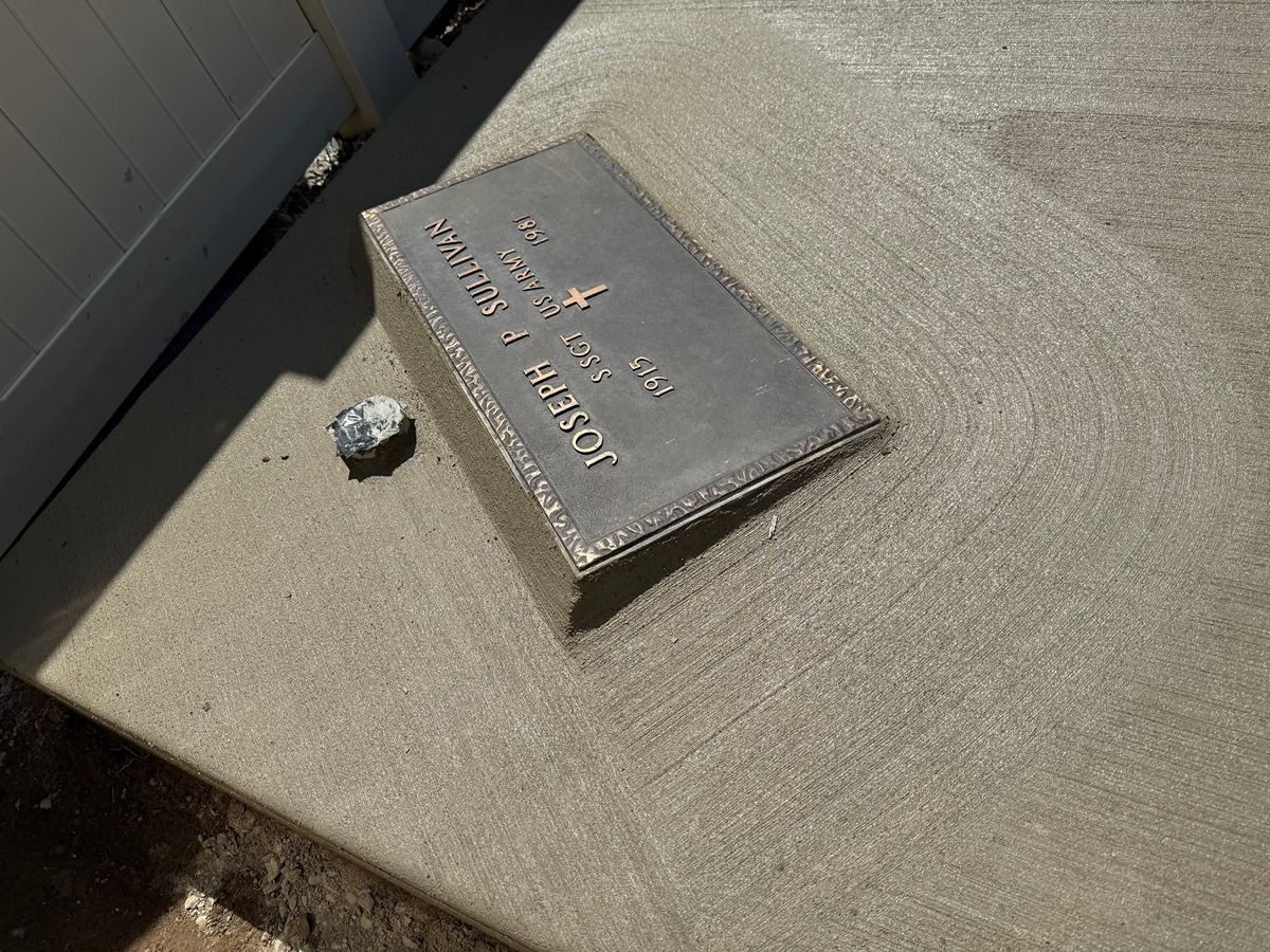 Small rectangular metal plaque on a concrete surface beside a wall, with a small rock nearby.