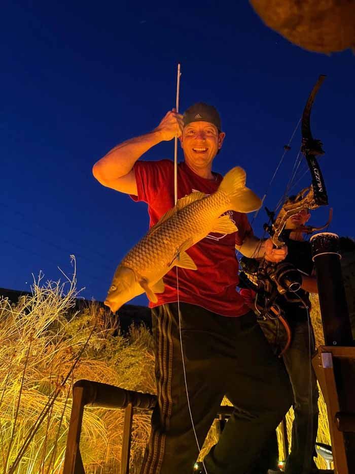Man holding bow and arrow, smiling, displaying a fish he has caught at dusk.