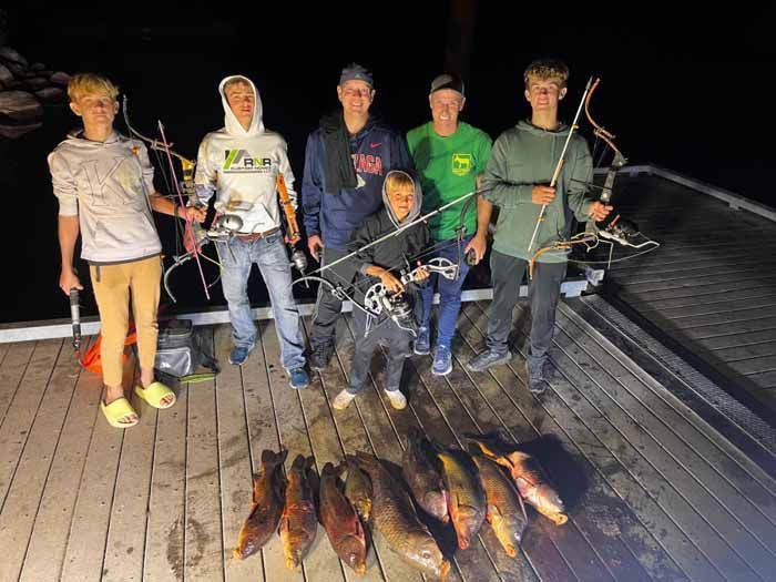 A group of young men are standing on a dock holding fishing rods and bows.