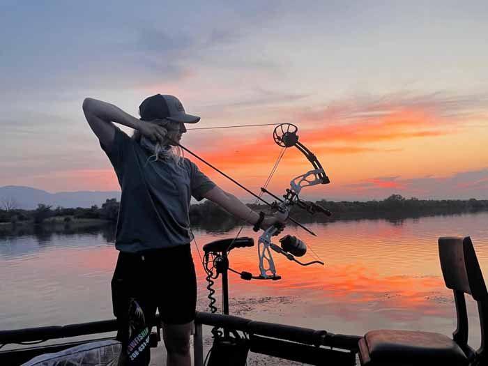 Person drawing bow on boat, aiming at sunset over water.