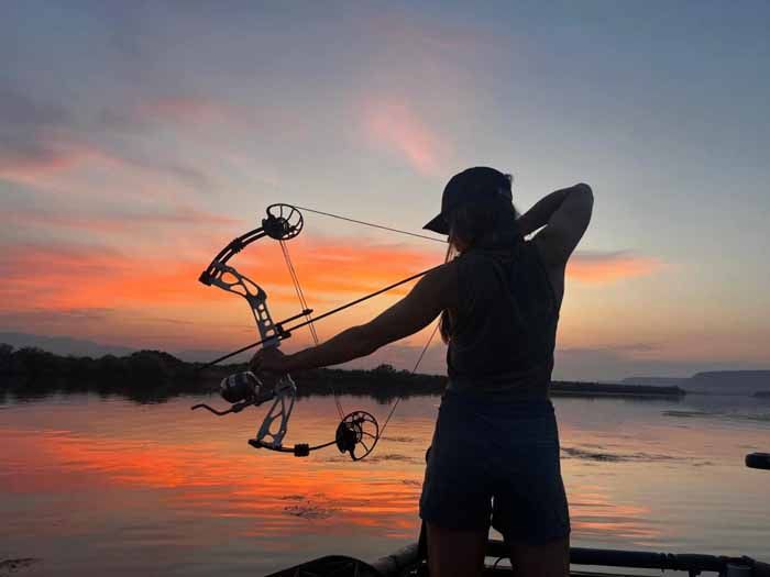 Person aiming a bow and arrow during a vibrant sunset over water, silhouetted.