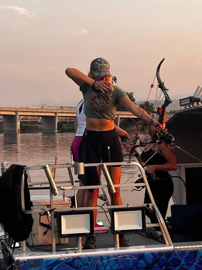 Woman aims bow and arrow from boat, river and bridge in the background. Sunset, arms outstretched.
