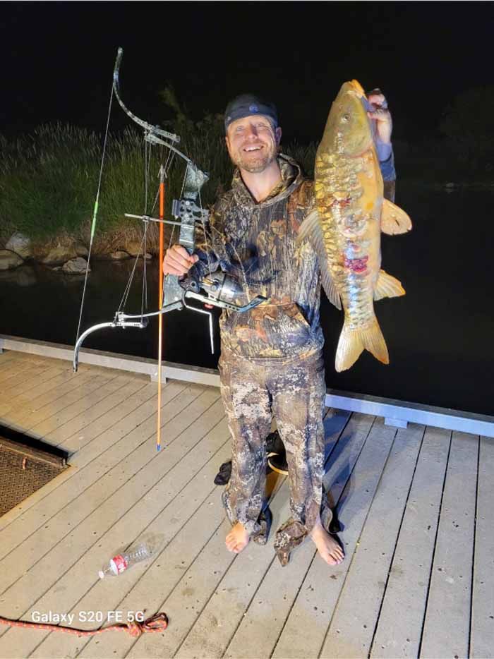 Man in camouflage holding a large fish and bow, standing on a dock at night.