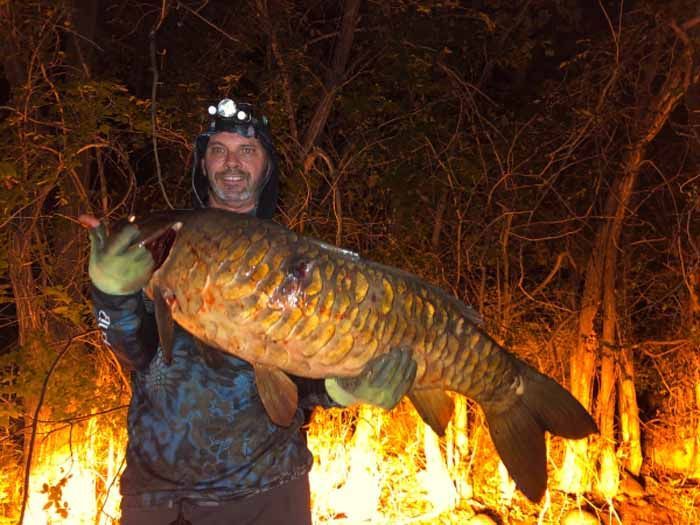 A man is holding a large fish in front of a fire