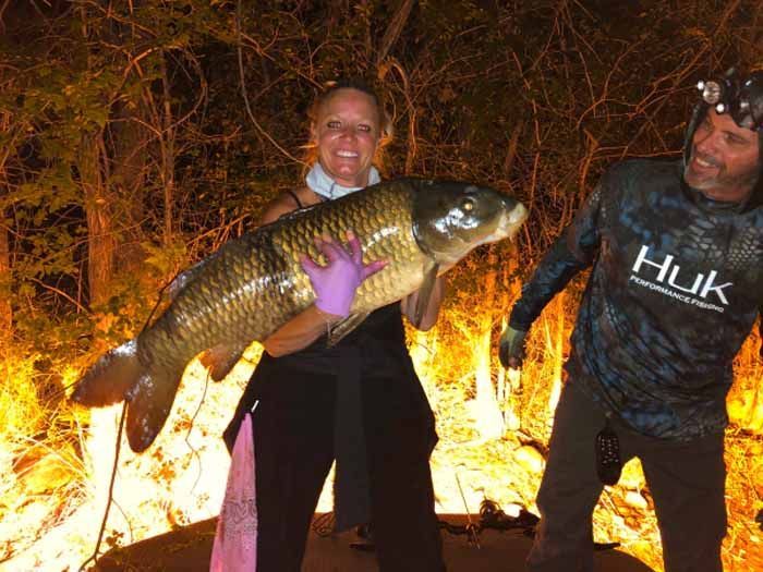 A woman is holding a large fish next to a man wearing a huk shirt