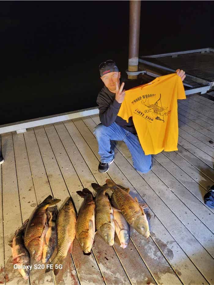 A man is kneeling down on a dock holding a t-shirt over a pile of fish.