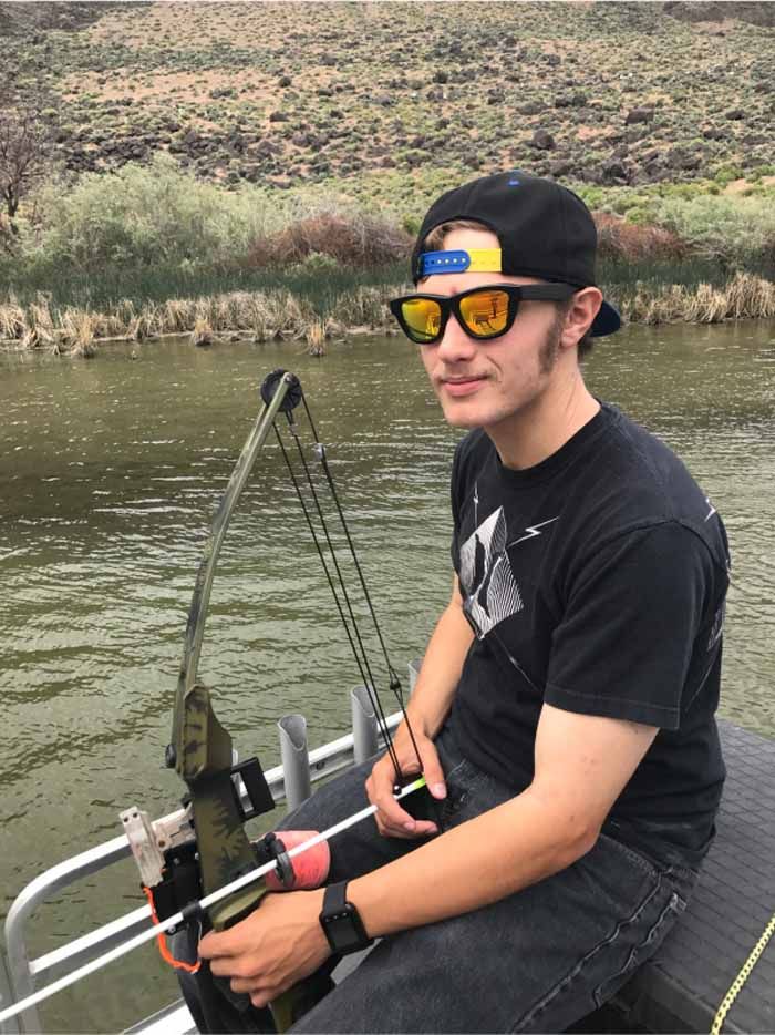 Man in sunglasses and hat on a boat, holding a bow and arrow, on a river.