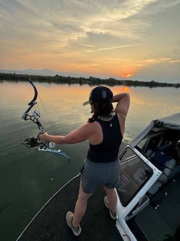 Person on a boat aiming a bow and arrow at sunset over water.