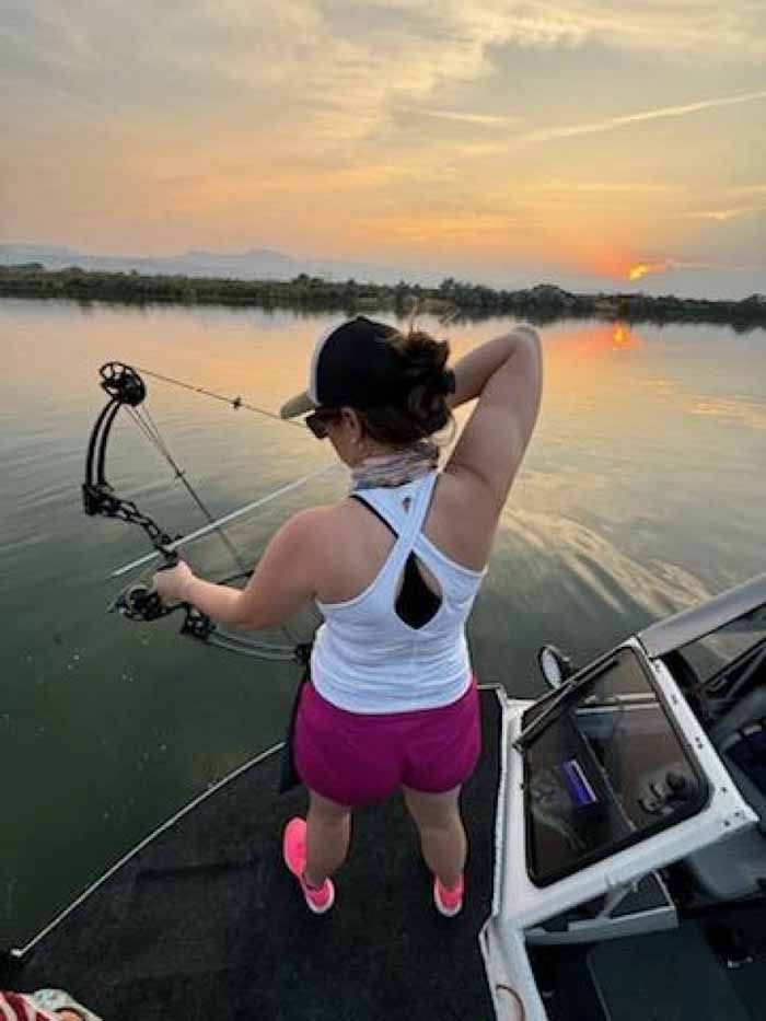 Woman on a boat draws bow and arrow against a sunset. She wears a cap, tank top, and shorts.