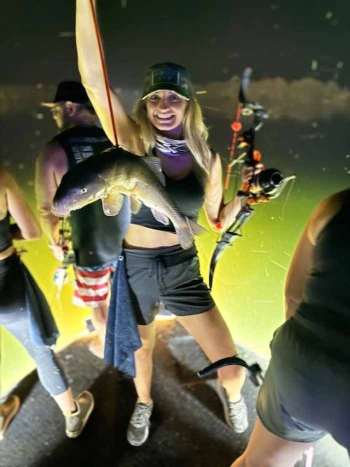 Woman holds a fish and bow, smiling. Other people stand around, night setting.