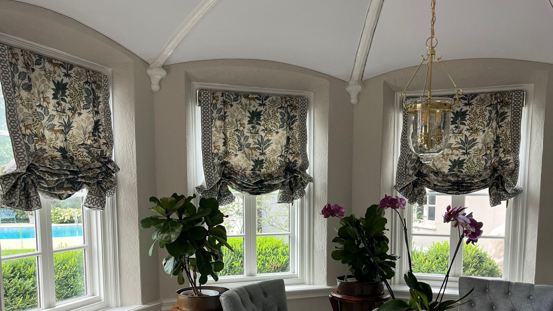 Bright sunroom with curved windows, patterned chairs, glass table, and potted plants.