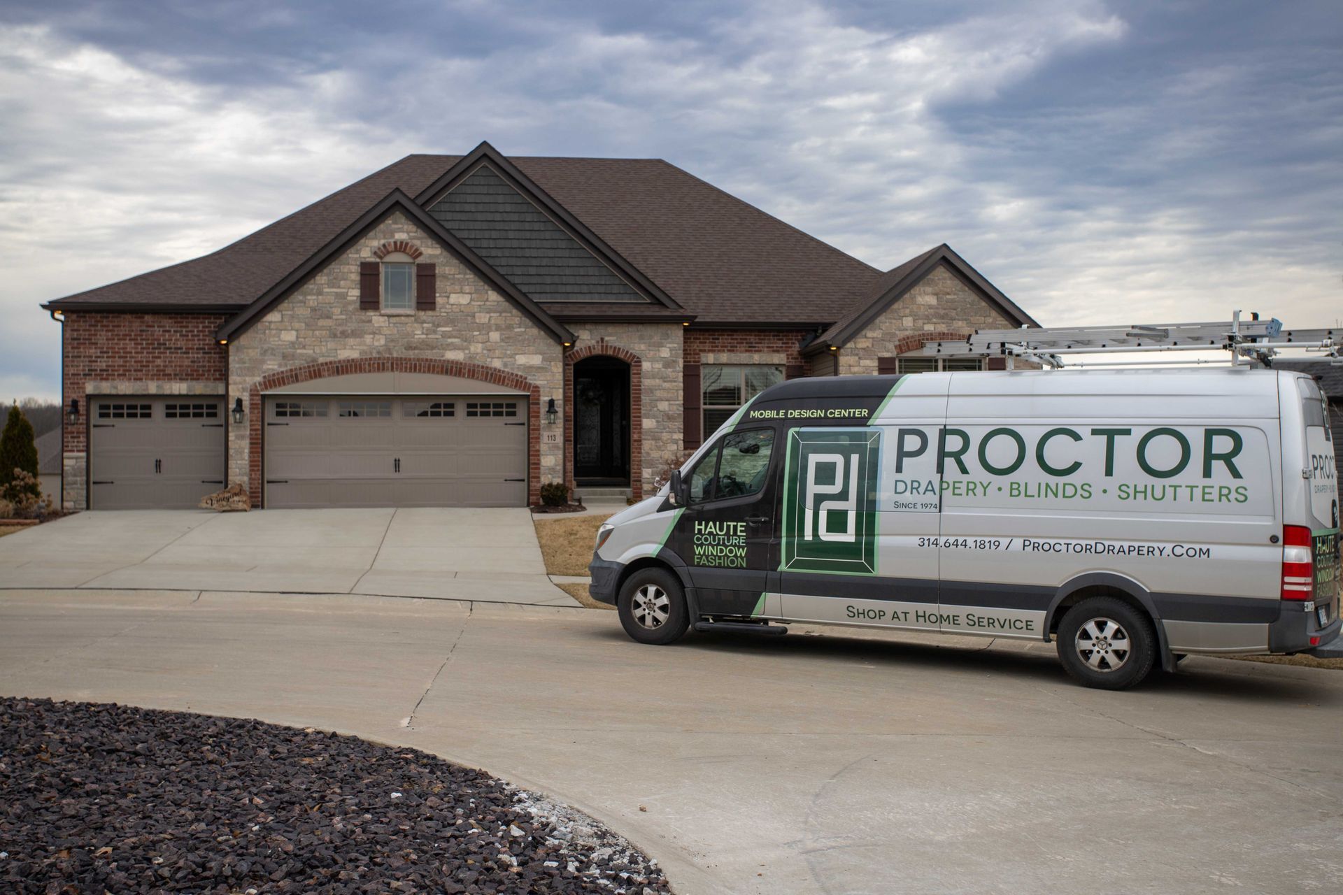 A silver Proctor Drapery, Blinds, & Shutters van parked in the driveway of a brick and stone suburban house.