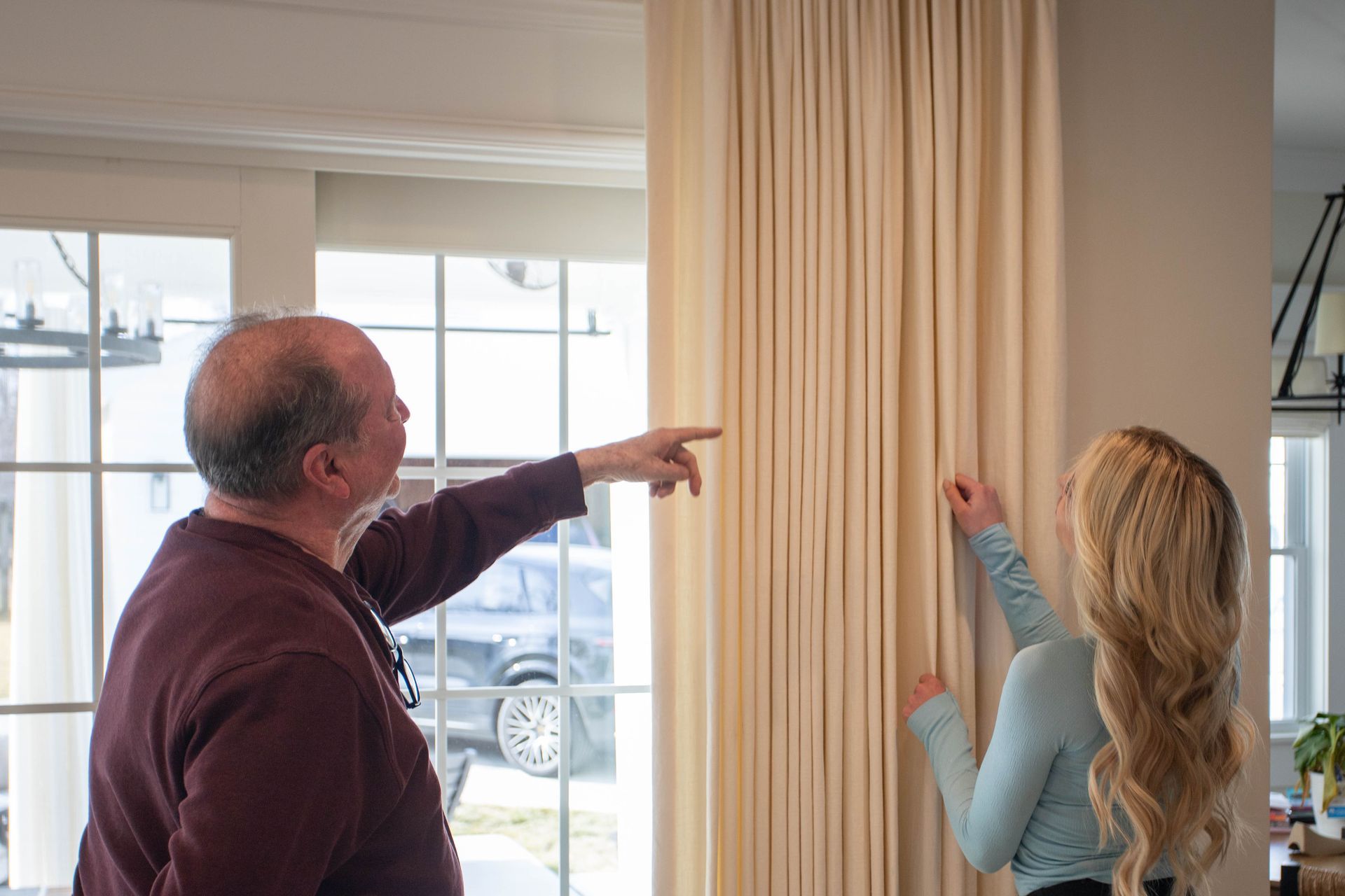 A man and woman inspect curtains, pointing. Light beige drapes hang by a window. Indoor setting.