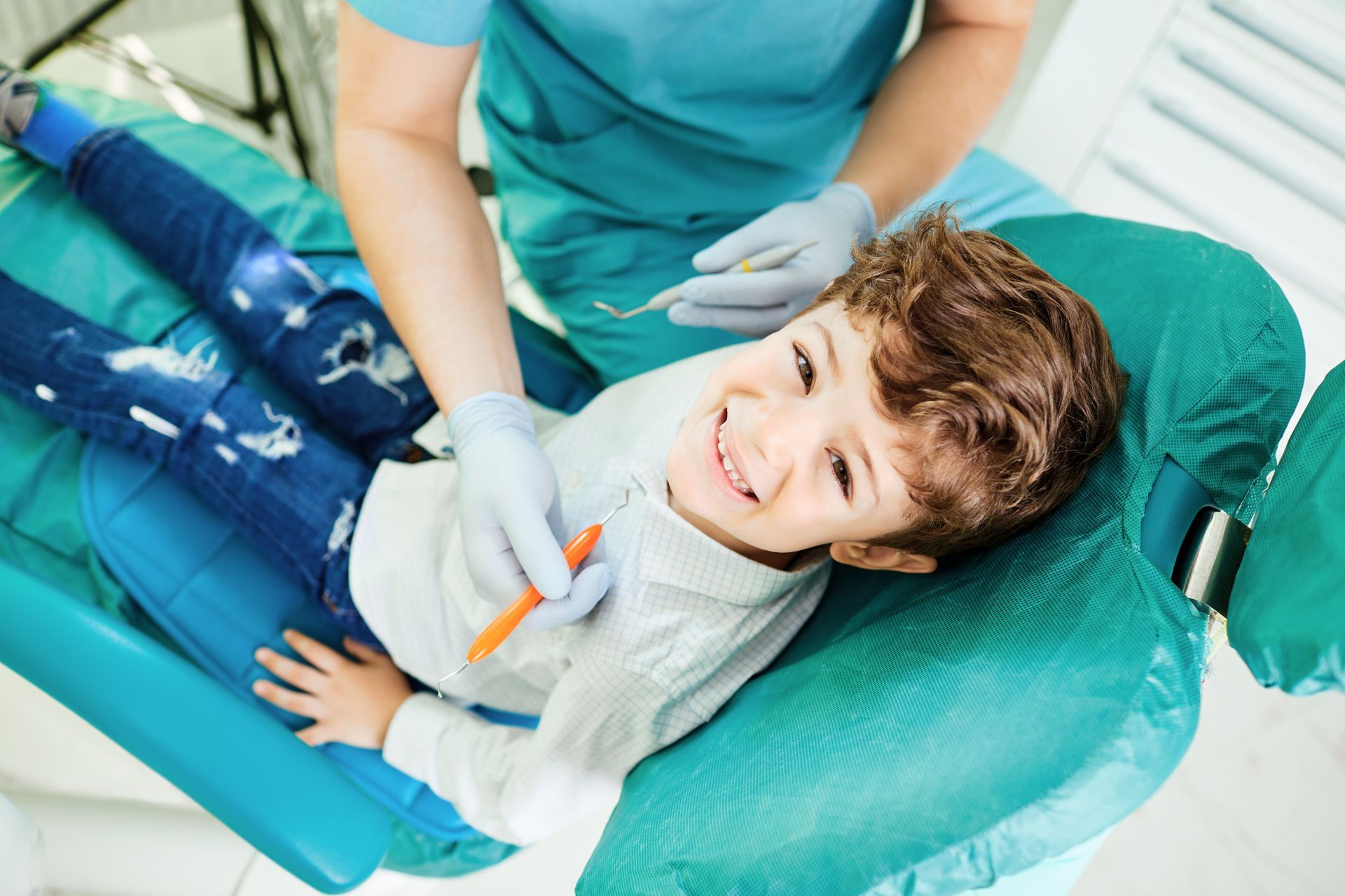 Smiling boy reclined in a teal chair while a gloved dentist performs a pediatric dentistry checkup.