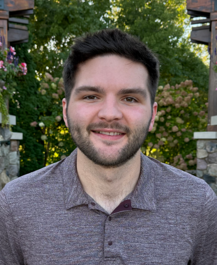 Man with short dark hair and a beard, smiling outdoors, wearing a maroon polo shirt.