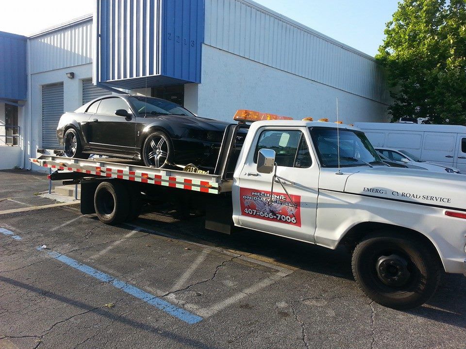 Tow truck carrying a black classic car parked beside a blue building | Mechanic On Duty CB LLC
