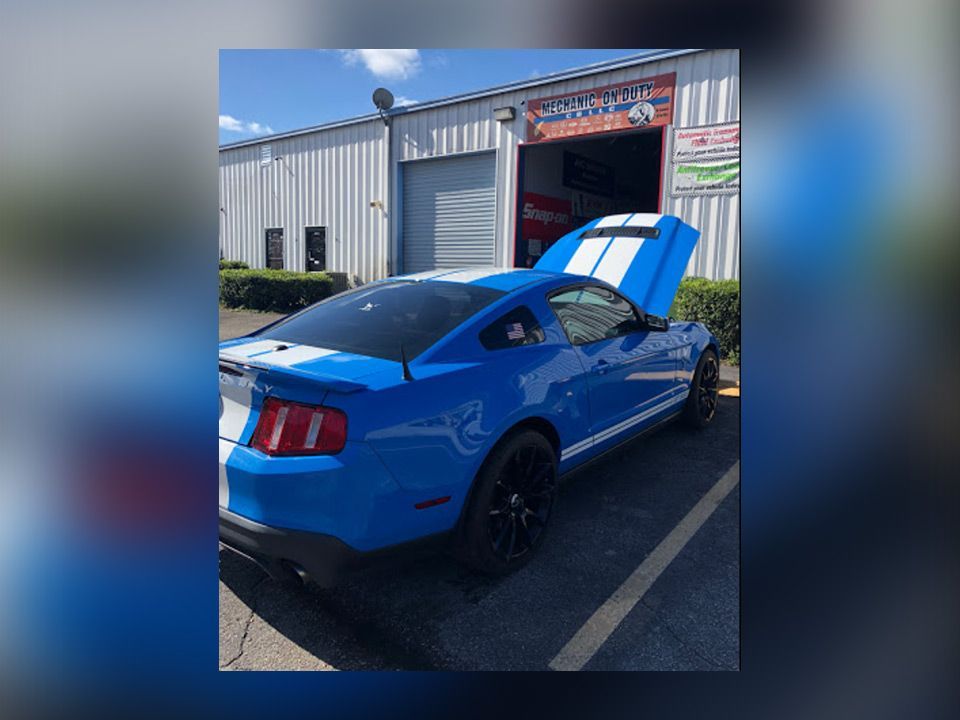 Blue sports car parked outside an auto shop with its hood open and rear lights visible | Mechanic On Duty CB LLC