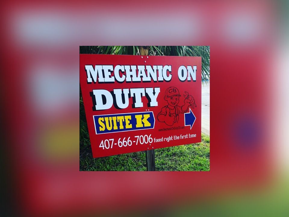 Red sign reading “Mechanic on Duty” for Suite K with phone number, on a grassy background. | Mechanic On Duty CB LLC