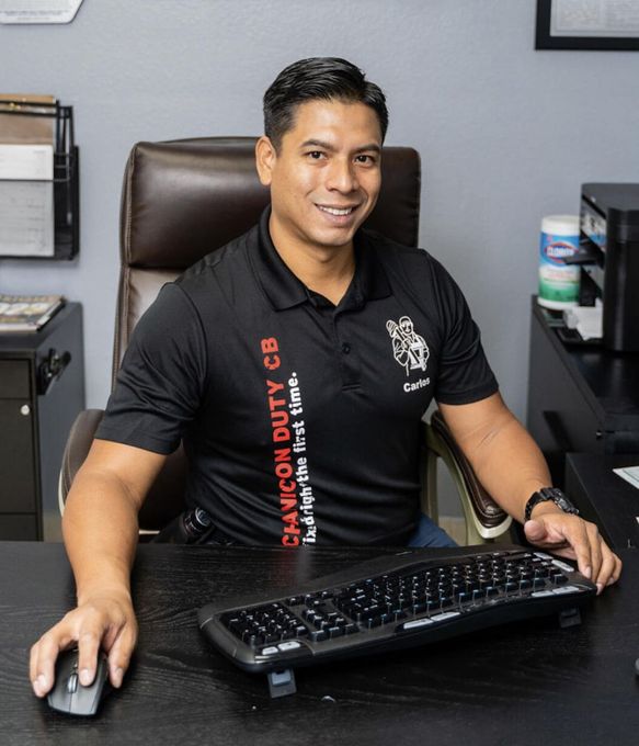 Person seated at an office desk using a mouse and keyboard, smiling in a black polo shirt. | Mechanic On Duty CB LLC