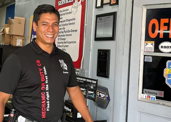 Smiling worker standing by a store entrance with posted signs and a payment window | Mechanic On Duty CB LLC