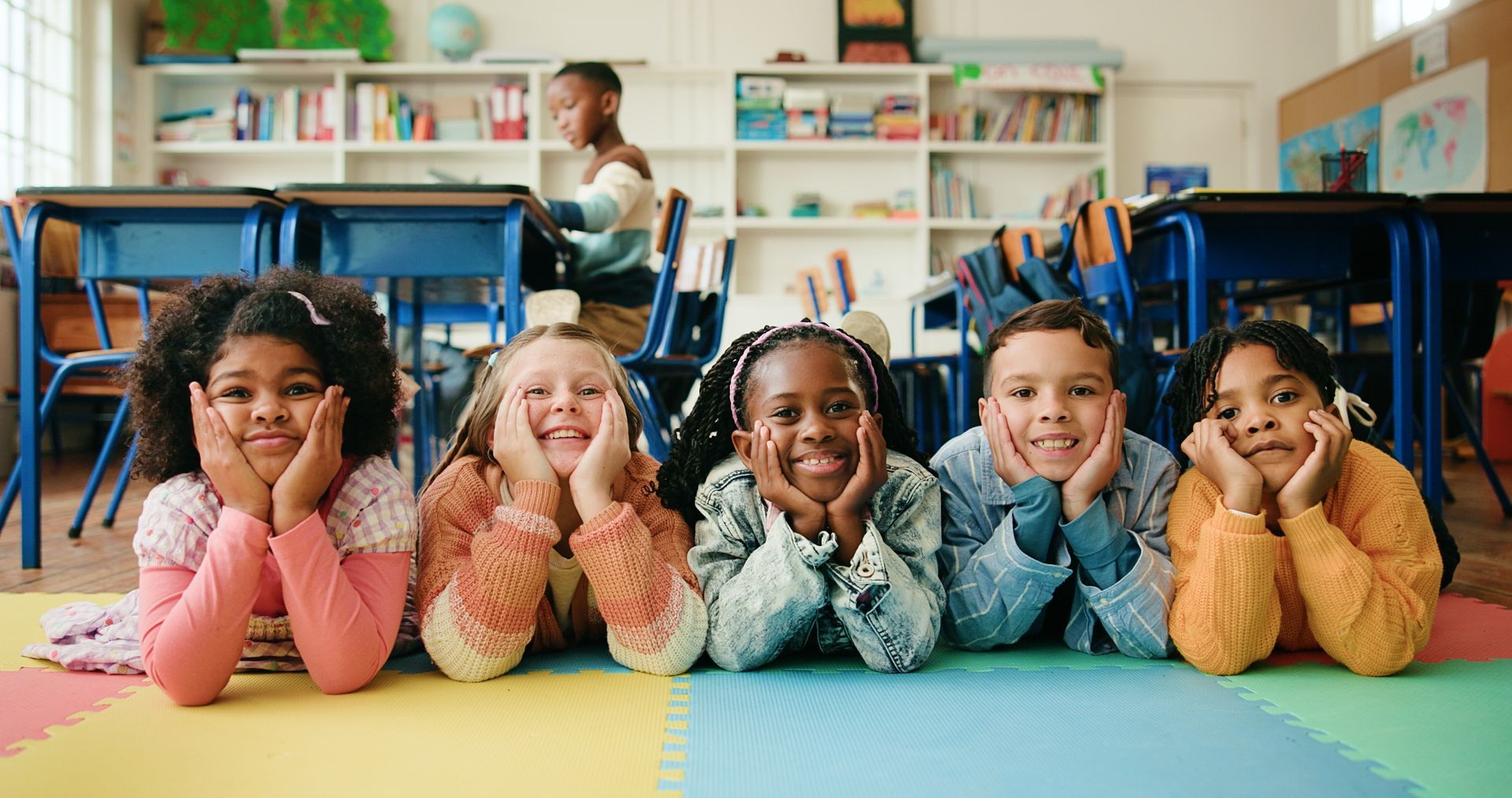 Five children lying on the floor in a classroom, smiling at the camera, with blue chairs and shelves behind them
