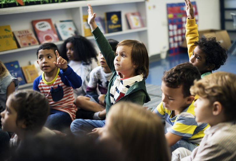 Children sitting on the floor in a classroom, raising their hands during a lesson