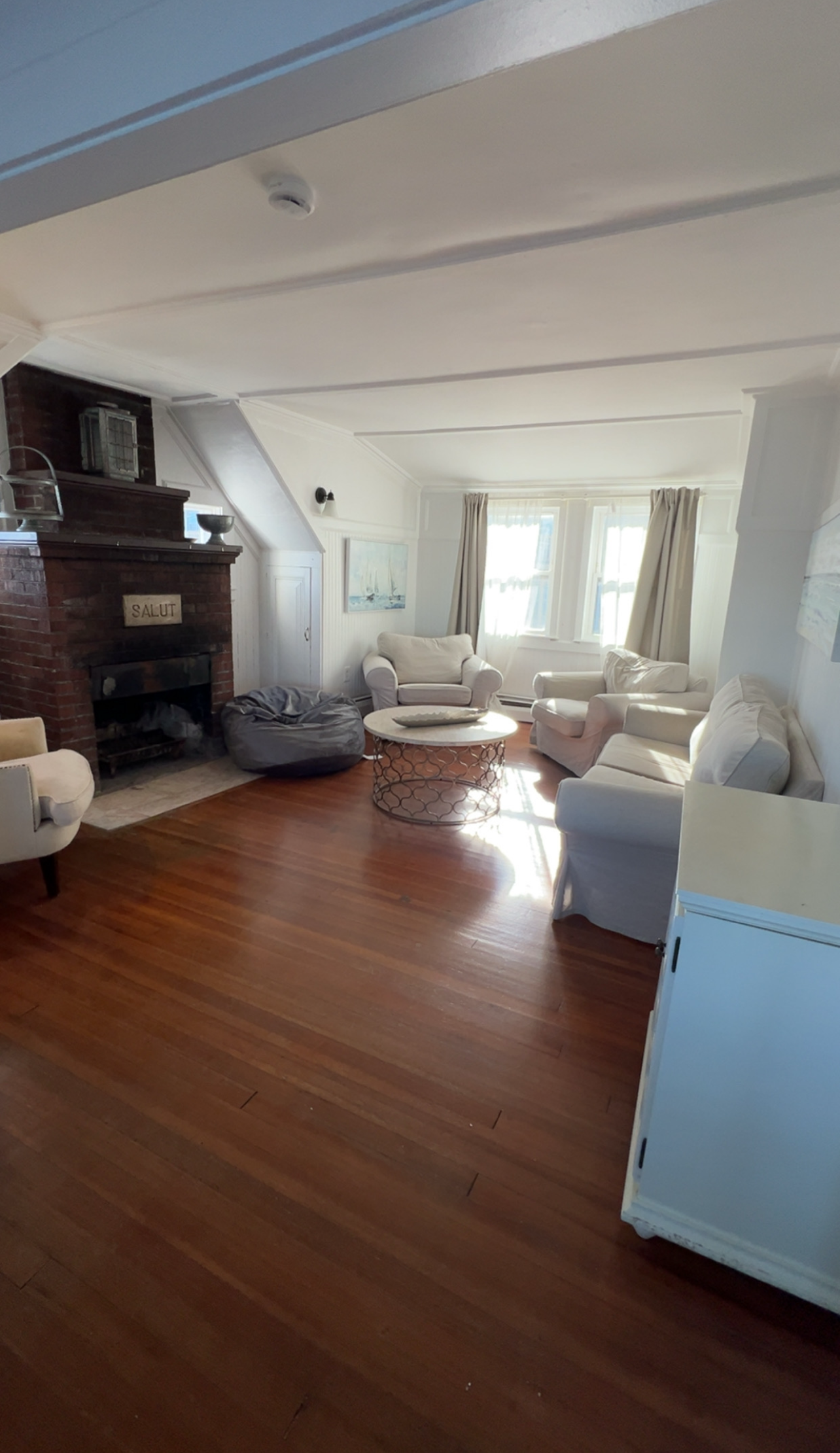 A cozy living room with a brick fireplace, hardwood floors, multiple white armchairs, and a round coffee table.