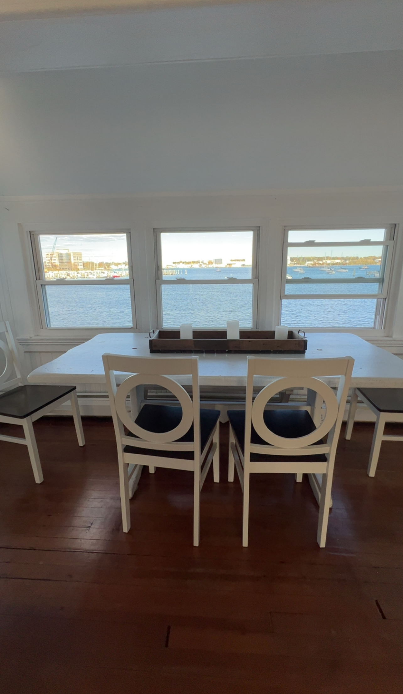 A dining table with four white chairs and a centerpiece sits in front of three large windows overlooking the ocean.