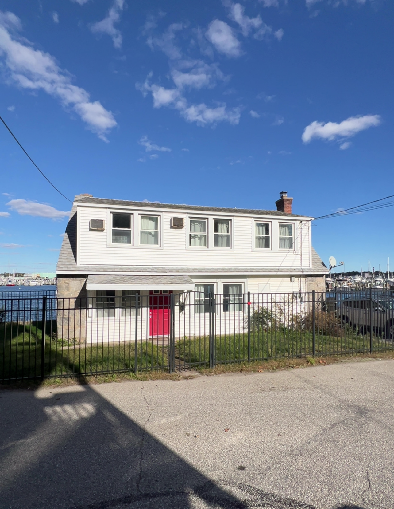 A two-story white house with a red front door behind a metal fence, located near a waterfront with boats in the distance.
