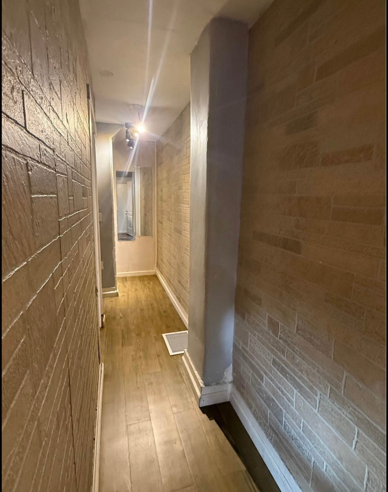 A narrow hallway with light wood flooring, beige textured brick walls, and a grey column leading to a bright doorway.