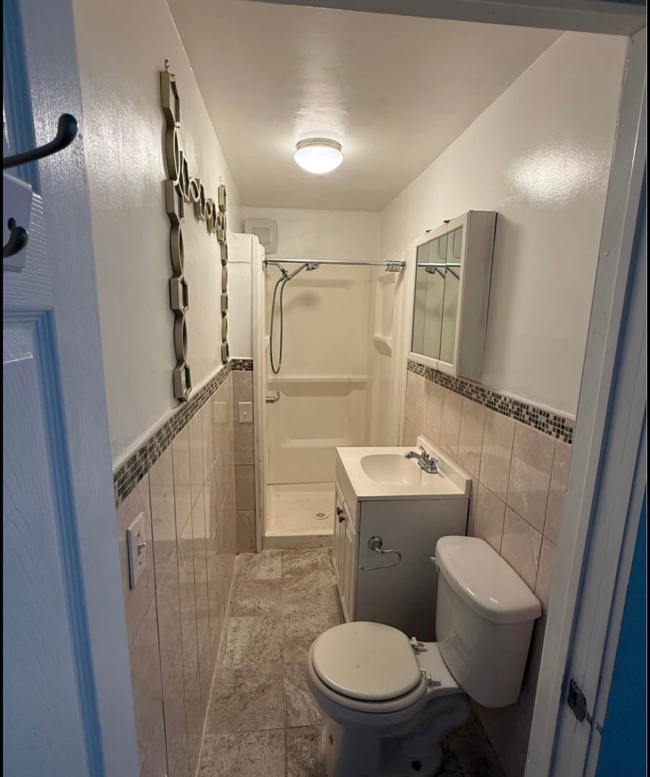 A narrow bathroom with white walls, tiled wainscoting, a pedestal sink, a toilet, and a walk-in shower at the far end.