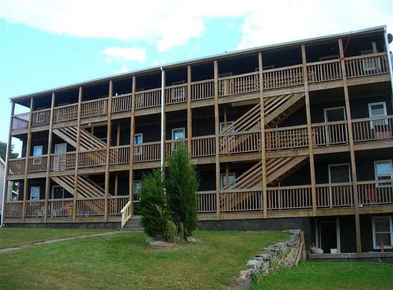 A three-story dark brown apartment building with exterior wooden balconies and staircases on a grassy lot.