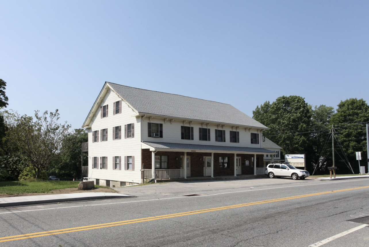 A three-story, white building with a gray roof and a front porch sits along a road under a clear blue sky.
