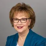 Smiling woman with short auburn hair and glasses, wearing a blue blazer against a gray background