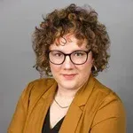 Portrait of a person with curly hair and glasses, wearing a mustard blazer against a gray background