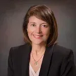 Professional headshot of a smiling woman in a black blazer against a gray background