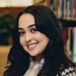 Smiling woman with long dark hair in a library, wearing a patterned top.