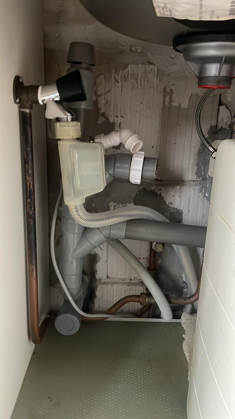 Leidingwerk onder de gootsteen met een waterkraan, een plastic sifon en grijze pvc-buizen tegen een kale bakstenen muur.