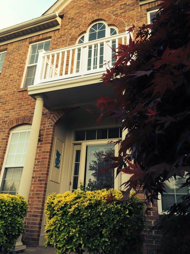 Brick townhouse entrance with white balcony, arched window, and green bushes in front